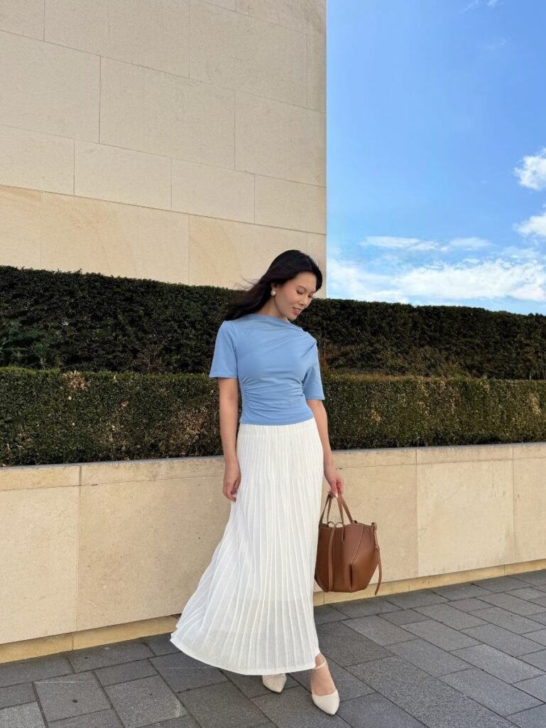 Woman wearinga  long pleated skirt, blue jersey blouse and brown handbag. 