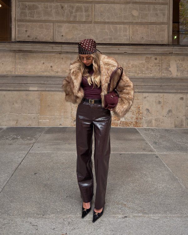 This image shows a woman in a brown faux fur jacket, a dark top, dark brown patent leather trousers, and a matching dark brown patent leather handbag and black heels