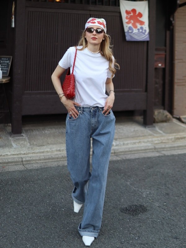 Woman wearing blue wide-leg jeans with a tucked-in white T-shirt, sneakers, sunglasses, and a crossbody bag for a casual street-style outfit.