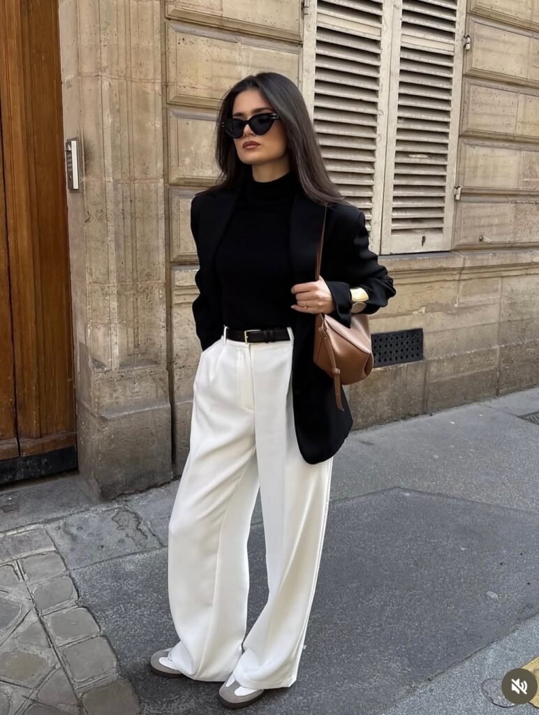 Woman wearing black wide-leg pants with a fitted black bodysuit, belt, sandals, sunglasses, blazer and clutch for a sleek day-to-night outfit idea.