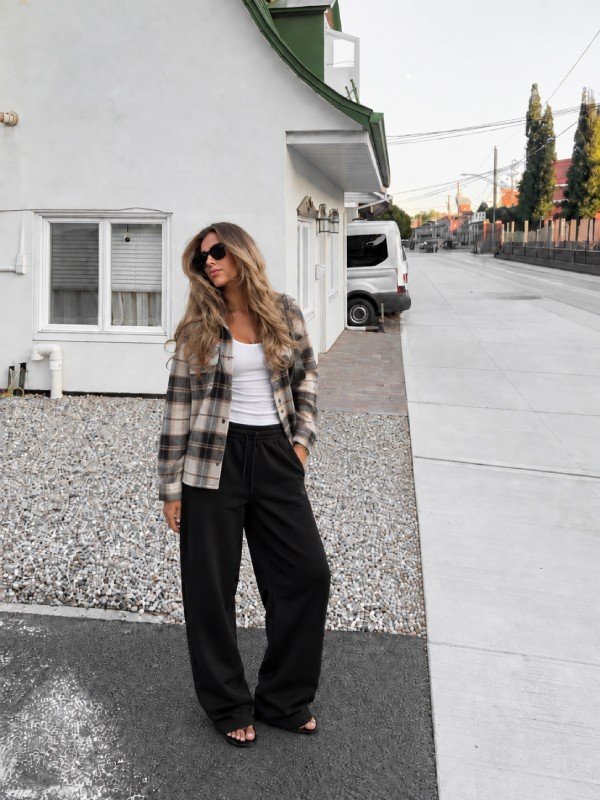 Blonde woman wearing a neutral plaid jacket over a white tank top with relaxed black sweatpants, black sandals, and sunglasses for a casual fall outfit.
