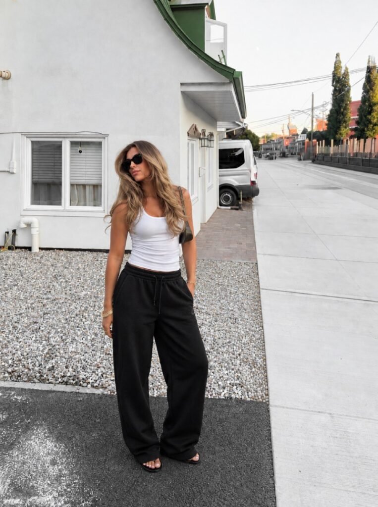 Blonde woman wearing a fitted white tank top with relaxed black sweatpants, sunglasses, and casual sandals for a simple minimalist outfit.