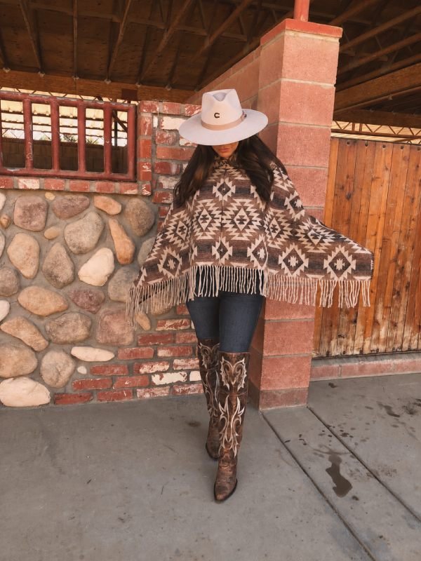 Woman wearing a cream poncho, jeans, a brown cowboy hat, and tall cowboy boots in a snowy landscape.